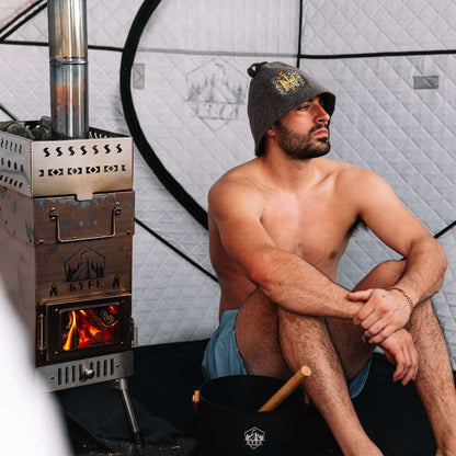 Man relaxing inside arctic gray KYFE portable sauna tent wearing sauna hat beside wood-burning stove with open fire – Finnish wood-fired sauna experience