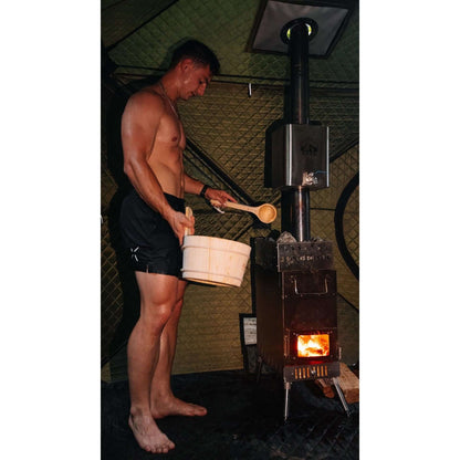 Man pouring water with wooden ladle and bucket onto KYFE wood-burning sauna stove with volcanic rocks inside forest green portable sauna tent – Finnish steam sauna experience